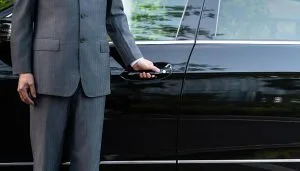 unrecognizable well dressed male chauffeur opening a car door A black limousine stands parked outside a large building, suggesting a nearby limo rental near Newark NJ